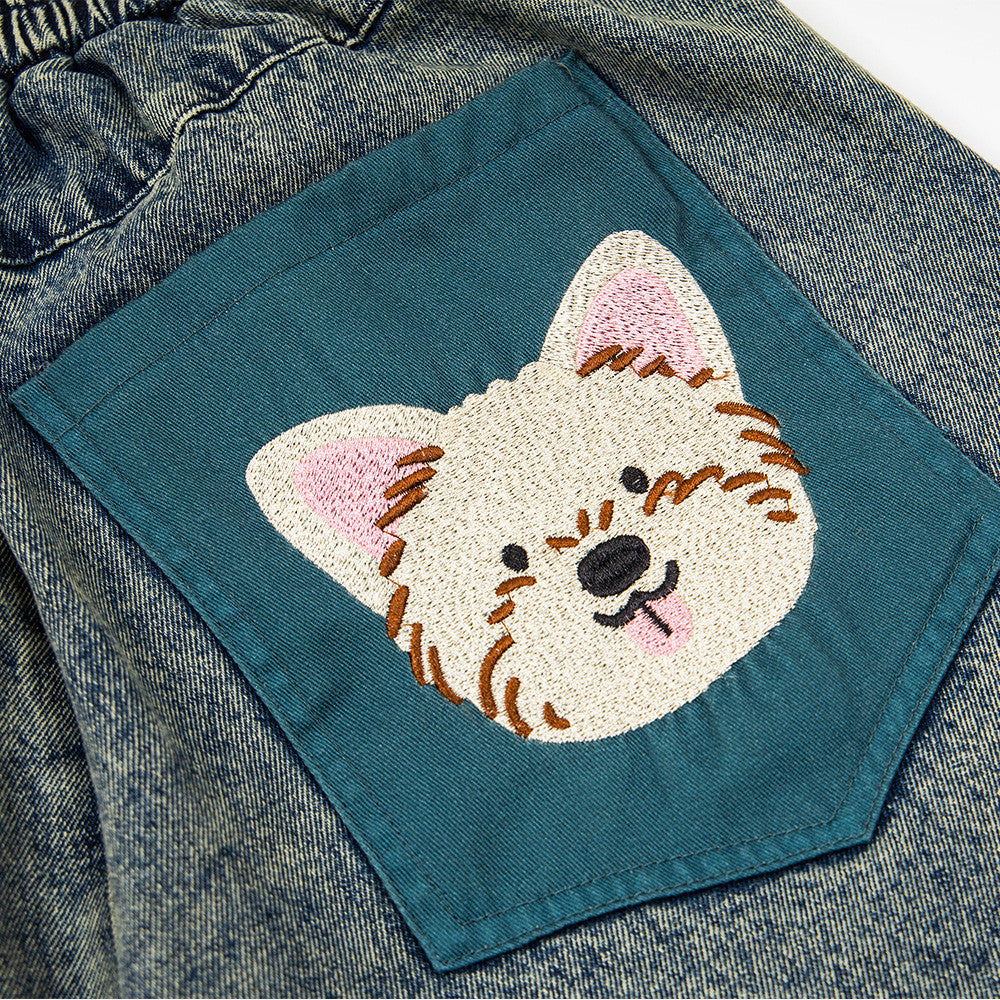 Denim shorts with teal pocket patch featuring a white embroidered dog face on distressed blue denim.