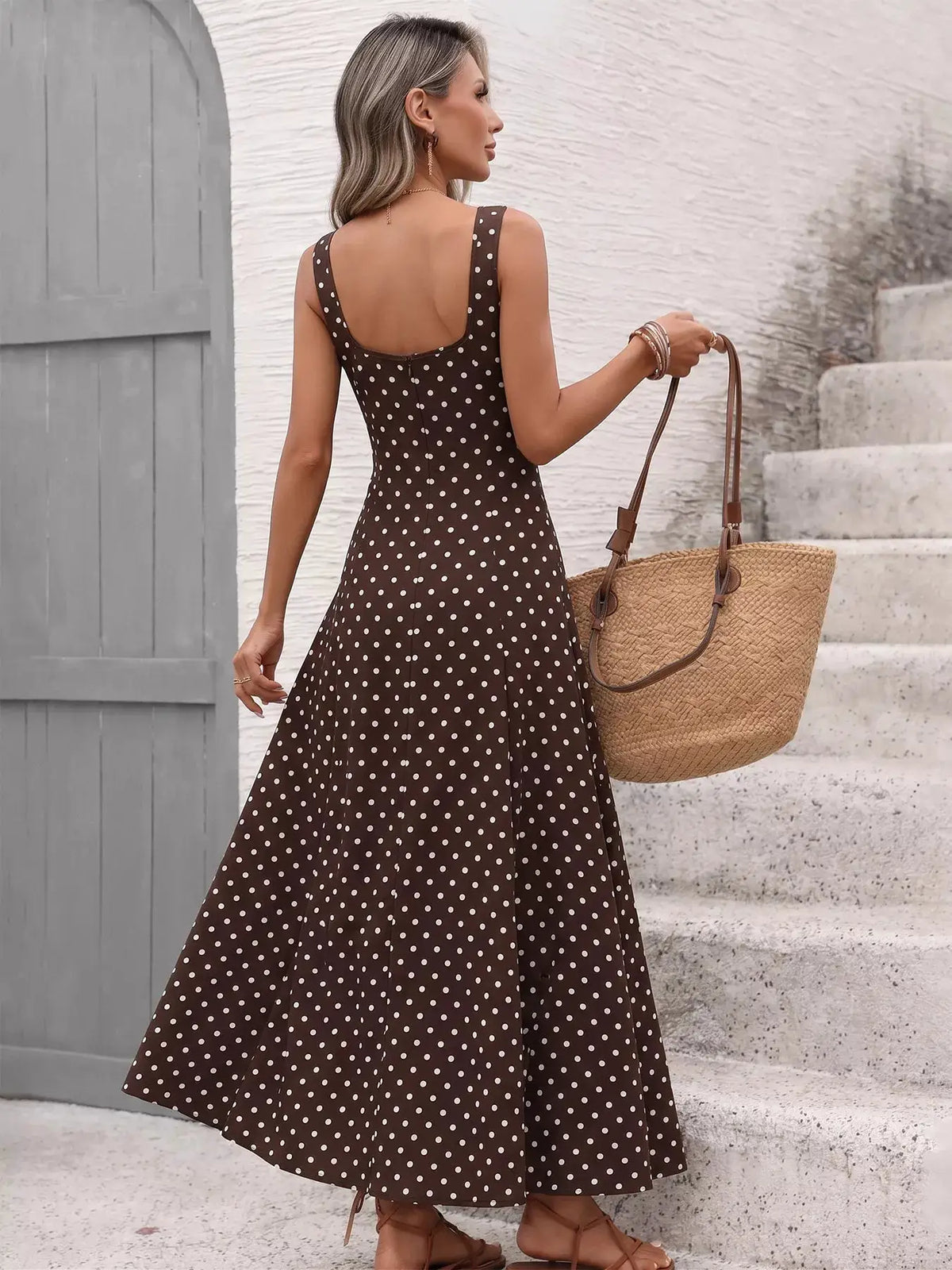 Polka dot sleeveless maxi dress in brown with white dots, paired with a straw tote bag