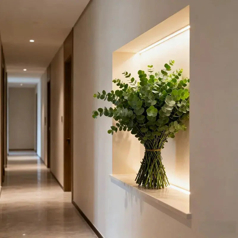 Artificial eucalyptus bouquet in a glass vase with green rounded leaves, displayed in a lit hallway niche.