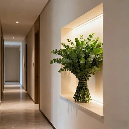 Artificial eucalyptus bouquet in a glass vase with green rounded leaves, displayed in a lit hallway niche.