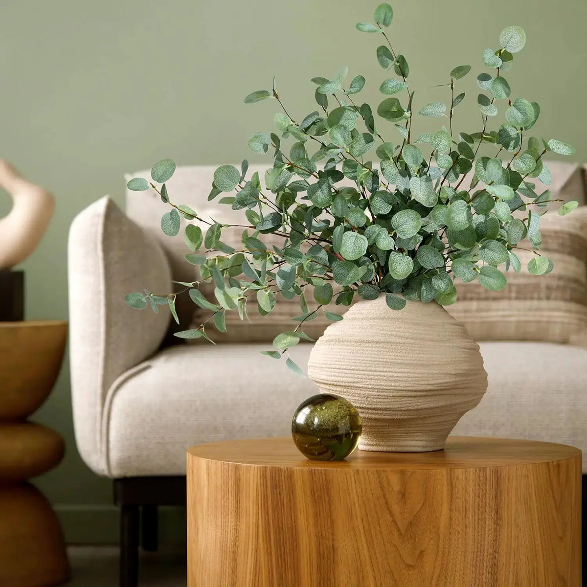 Artificial eucalyptus stems bouquet in a textured beige vase on a wooden table